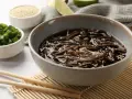 Miseczka z makaronem soba w sosie, posypana sezamem. Obok miseczki z posiekanym szczypiorkiem i sezamem. Zdrowy posiłek, mało makaron soba kcal.