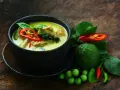 Aromaticzne zielone curry z kawałkami mięsa, papryczką chili, zielonym pieprzem i liśćmi bazylii. Obok miski świeże składniki: limonka kaffir, pomidorki i papryczki chili.