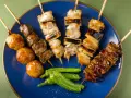Różnorodne szaszłyki kurczak yakitori: mięso, wątróbka, kulki mięsne i warzywa na niebieskim talerzu.