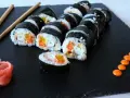 Roladki sushi z surimi, ryżem i warzywami na czarnej desce, obok pałeczki i różowy imbir.