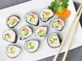 Talerz z rolkami sushi z awokado, ogórkiem i marchewką. Obok pałeczki i dekoracja z marchewki z wasabi. Idealna ilość octu do sushi podkreśla smak.