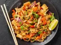 Pyszny pad thai z warzywami: makaron ryżowy, tofu, papryka, cebula i groszek. Podany z limonką i pałeczkami.