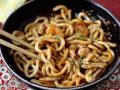 Pyszne udon z krewetkami w sosie, posypane sezamem i natką pietruszki. Danie w czarnej misce, gotowe do jedzenia pałeczkami.