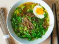 A miseczce aromatyczny rosół japoński z makaronem soba, jajkiem na pół i zieloną cebulką. Obok leżą pałeczki i surowy makaron.