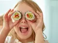 Uśmiechnięta dziewczynka trzyma kawałki sushi dla dzieci przed oczami, udając, że ma rolki zamiast oczu.