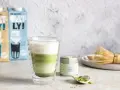 Warstwowy napój matcha z mlekiem kokosowym w szklance z podwójnymi ściankami. Obok puszka z matchą i bambusowa miotełka.
