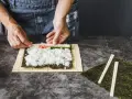 Ręce układają składniki do zwijania sushi na macie bambusowej. Ryż, paluszki krabowe i ogórek czekają na zawinięcie.