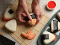 Ręce formują onigiri z krewetkami, tworząc pyszne japońskie jedzenie. Obok miska ryżu, nori i talerz gotowych przysmaków.