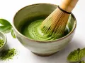 Tradycyjne matcha przygotowanie: bambusowa miotełka miesza zielony proszek z wodą w miseczce, tworząc piankę. Obok mała miseczka z proszkiem i drewniana łyżeczka.