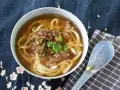 Gorąca zupa udon z wołowiną i szczypiorkiem, ozdobiona kolendrą, w misce na tle z motywem kwiatowym. Obok leży niebieska łyżka.