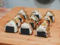 Rząd trójkątnych onigiri z kurczakiem, posypanych sezamem i przyprawami, ułożonych na drewnianej desce.