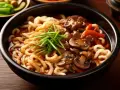 Miseczka pełna grubego makaronu udon w sosie teriyaki z kawałkami wołowiny, pieczarkami i szczypiorkiem.