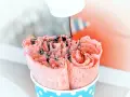 Różowe lody tajskie, zwinięte w rolki, polane czekoladą. Widać składniki, które tworzą ten deser.
