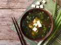 Aksamitne miso z tofu, wodorostami i szczypiorkiem, podane z pałeczkami na drewnianym blacie.