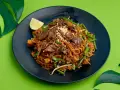 Pyszne pad thai z wołowiną, fasolką szparagową i orzeszkami na czarnym talerzu, ozdobione ćwiartką cytryny i liśćmi monstery.