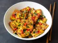 Chrupiące tofu w tempurze, posypane sezamem, chili i natką pietruszki, podane w białej misce z pałeczkami.