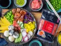 Kolorowy bowl z kawałkami glazurowanego lososia, ryżem, awokado, mango, ogórkiem, rzodkiewką, edamame i marynowanym imbirem. Obok opakowanie różowego imbiru.