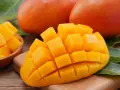 Dojrzałe mango, pokrojone w kostkę, prezentuje swoje soczyste, pomarańczowe wnętrze. Obok leżą całe owoce i liście.