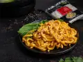Grube, pomarańczowe kluski biang biang w sosie, posypane sezamem i chili, z zielonymi liśćmi bok choy.