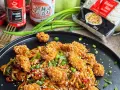 Pyszny makaron udon z kurczakiem w panierce, posypany sezamem i szczypiorkiem. Obok sosy i opakowanie makaronu.