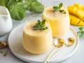 Dwa pyszne mango lassi z pulpy, udekorowane miętą i kardamonem, obok świeżego mango.