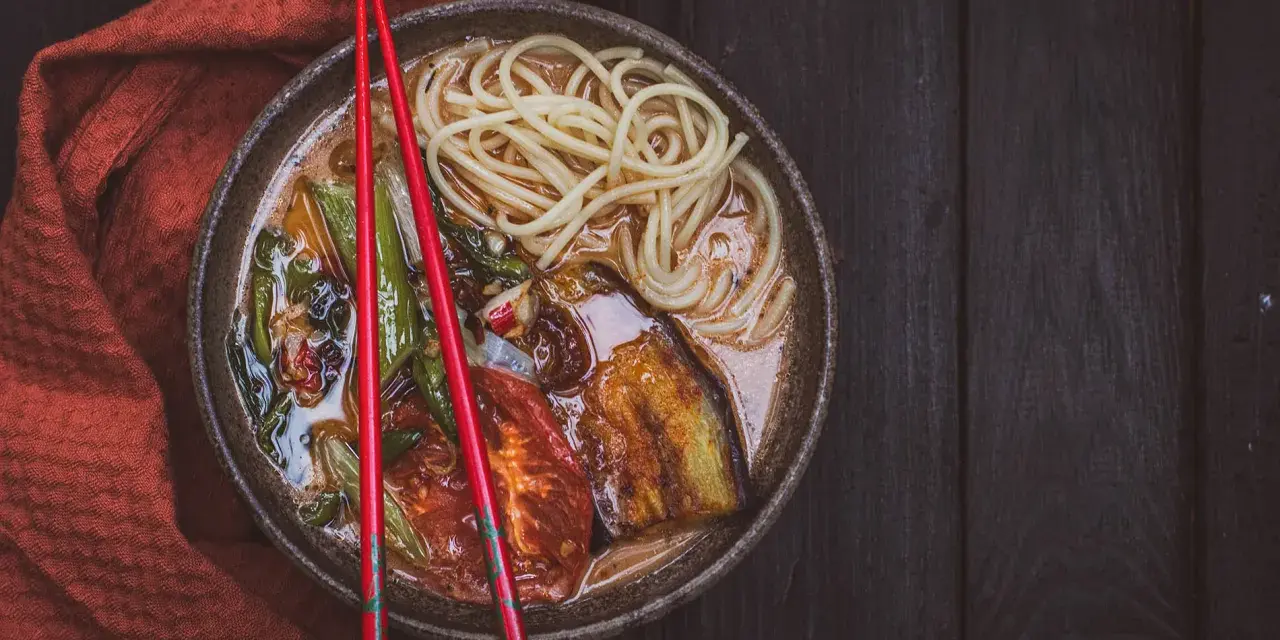 A miseczce z aromatycznym ramen wegetariańskim, pełnej makaronu, grillowanego bakłażana i pomidorów, spoczywają czerwone pałeczki.