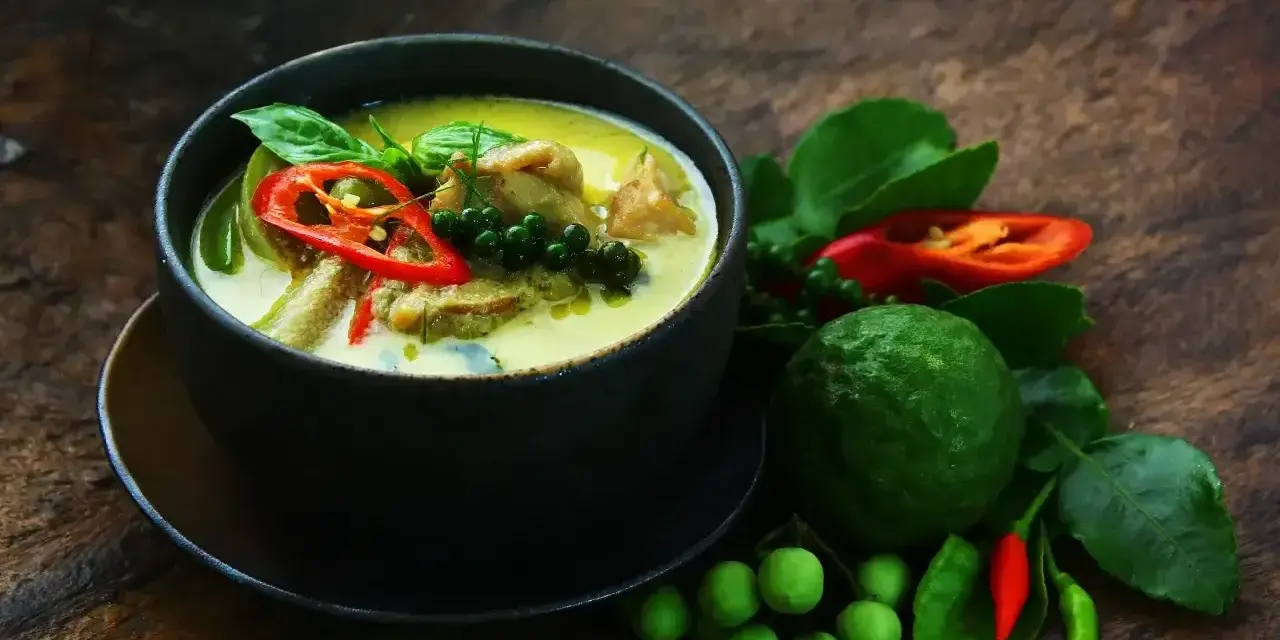 Aromaticzne zielone curry z kawałkami mięsa, papryczką chili, zielonym pieprzem i liśćmi bazylii. Obok miski świeże składniki: limonka kaffir, pomidorki i papryczki chili.