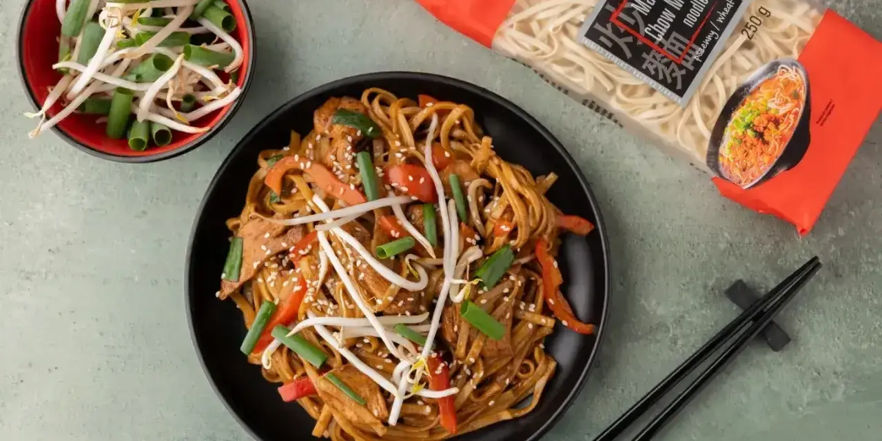 Pyszny azjatycki obiad z makaronem chow mein, warzywami i kiełkami fasoli. Obok talerza paczka makaronu i pałeczki.