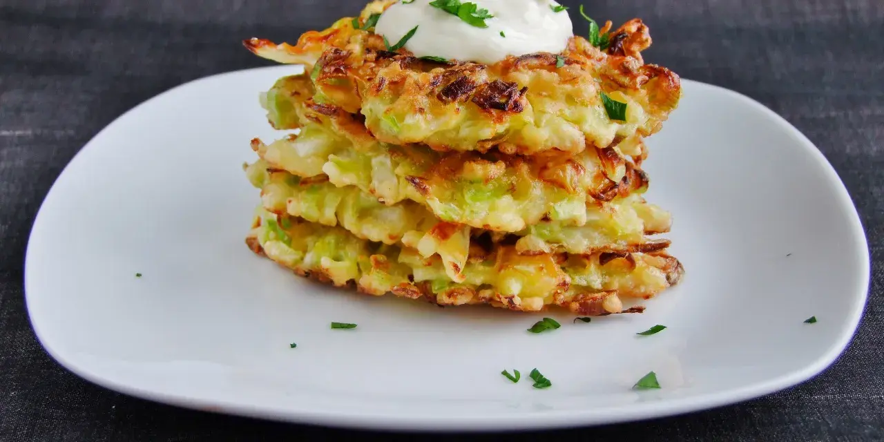 Stos placków okonomiyaki, polanych śmietaną i posypanych natką pietruszki, na białym talerzu.