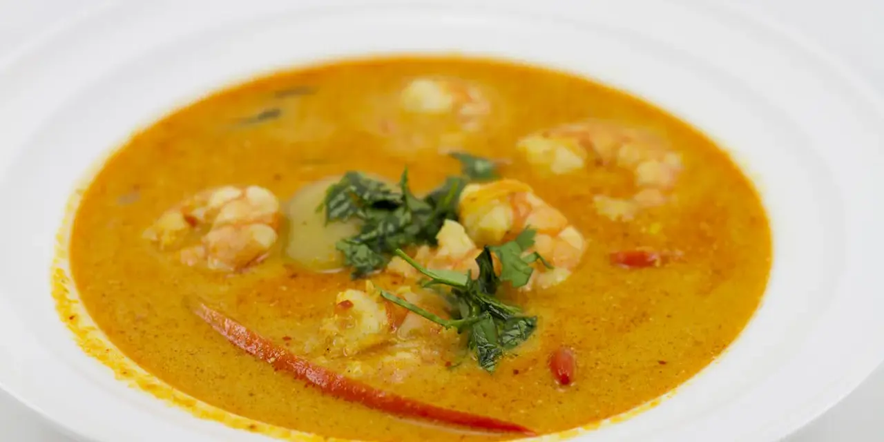 Aksamitna zupa tom yum z krewetkami, świeżą kolendrą i kawałkami chili na białym talerzu.