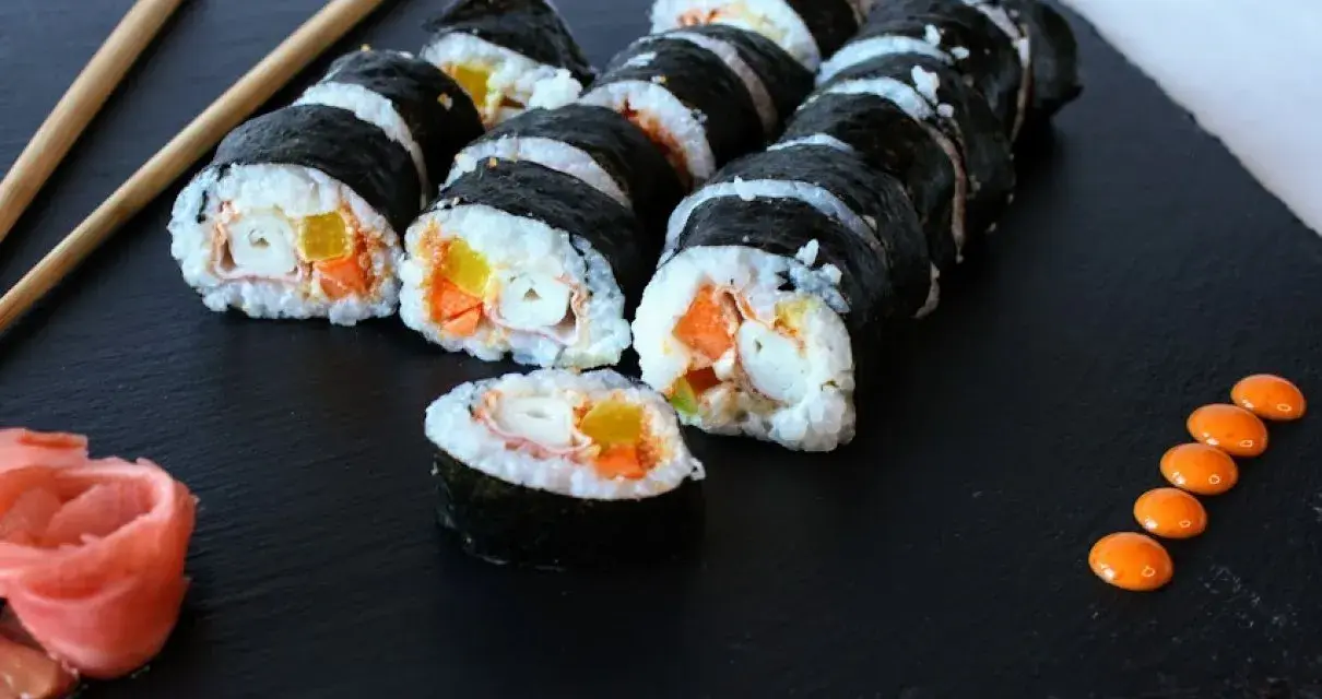 Roladki sushi z surimi, ryżem i warzywami na czarnej desce, obok pałeczki i różowy imbir.