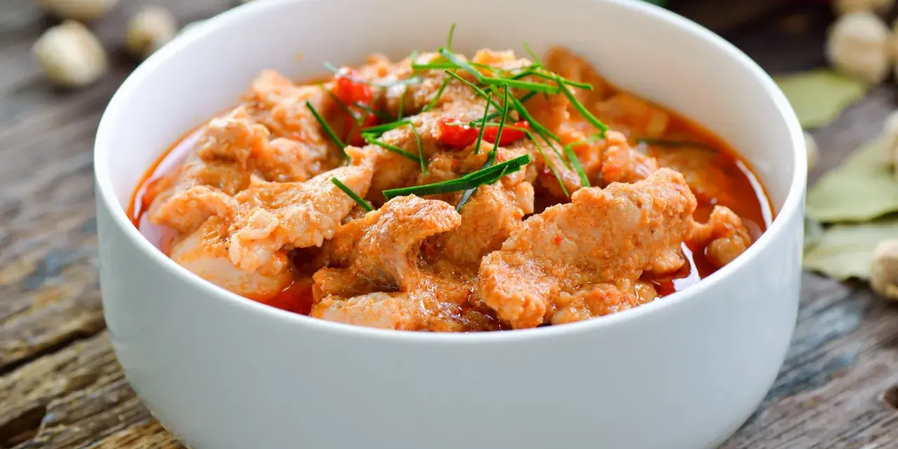 Pyszne wieprzowina w sosie curry, udekorowana szczypiorkiem i papryczką chili, podana w białej misce na drewnianym stole.