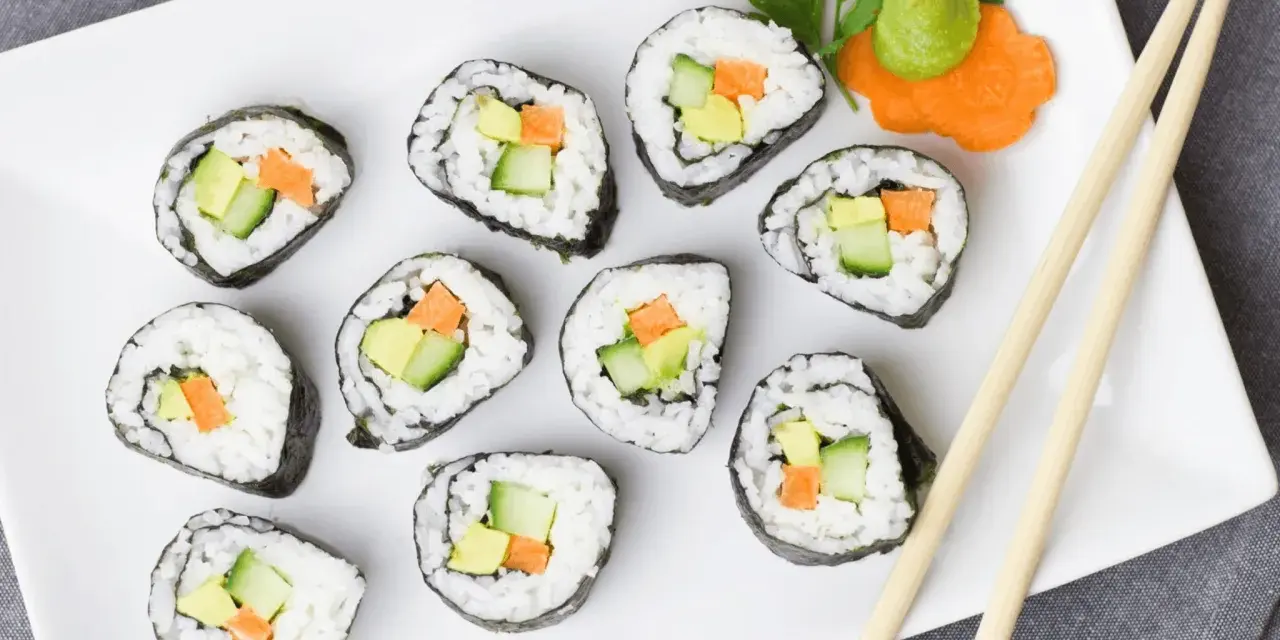 Talerz z rolkami sushi z awokado, ogórkiem i marchewką. Obok pałeczki i dekoracja z marchewki z wasabi. Idealna ilość octu do sushi podkreśla smak.