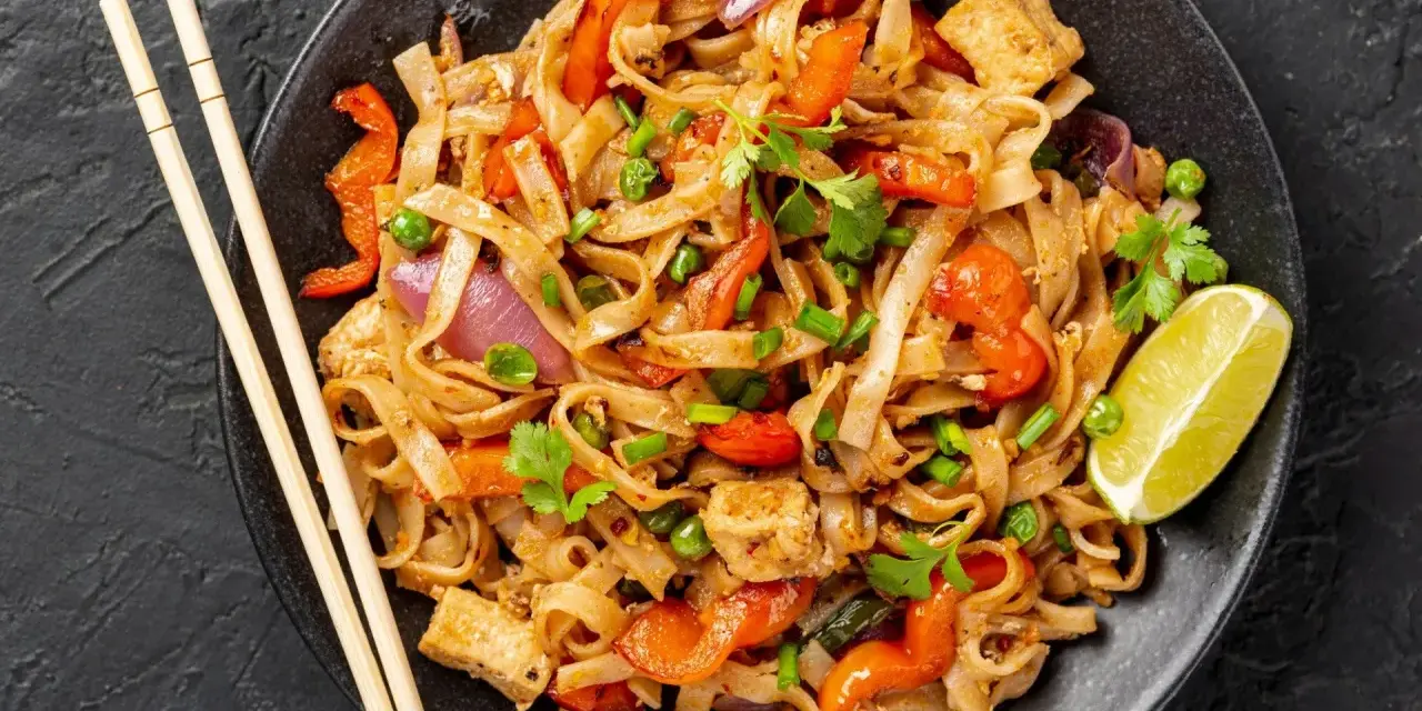 Pyszny pad thai z warzywami: makaron ryżowy, tofu, papryka, cebula i groszek. Podany z limonką i pałeczkami.