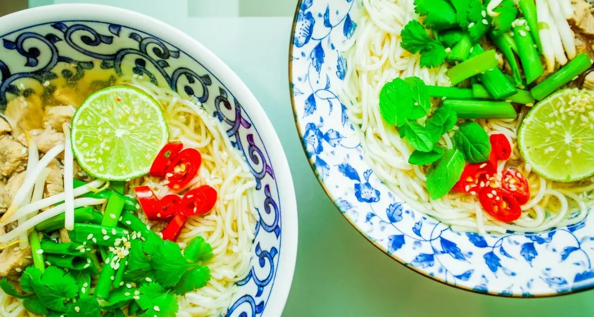 Dwie miseczki aromatycznej zupy pho z makaronem, mięsem, kiełkami, limonką, chili i świeżymi ziołami.