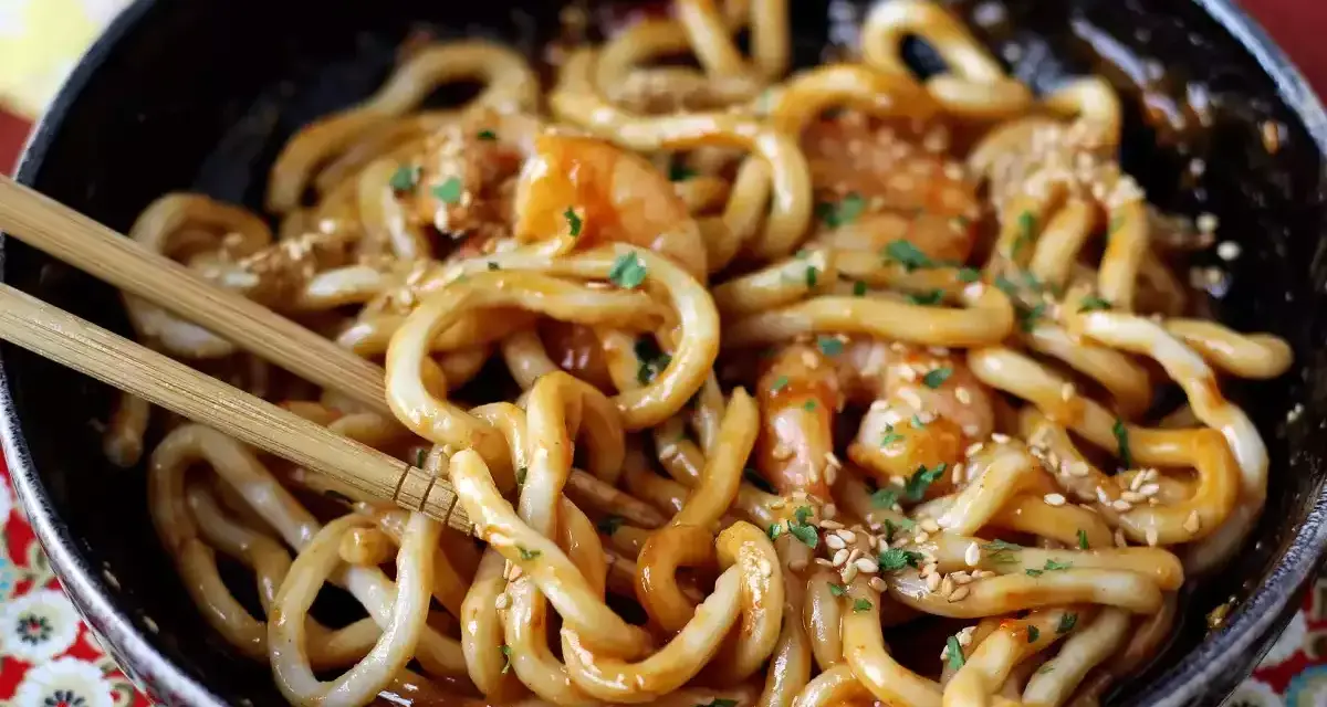 Pyszne udon z krewetkami w sosie, posypane sezamem i natką pietruszki. Danie w czarnej misce, gotowe do jedzenia pałeczkami.