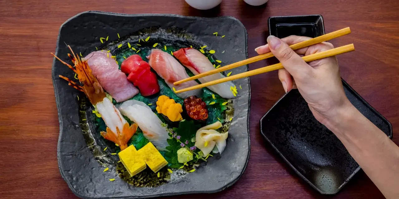 Wykwintne jedzenie w Japonii: ręka z pałeczkami sięga po świeże sushi i sashimi, obok sos i sake.
