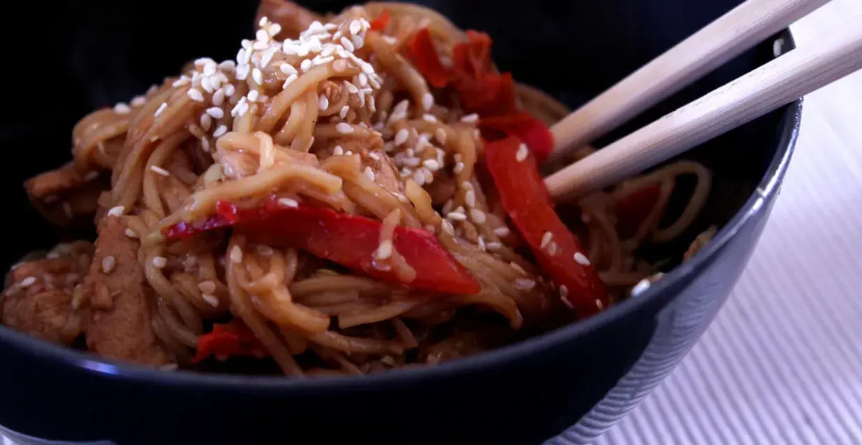 Miseczka pełna aromatycznego makaronu teriyaki z kawałkami kurczaka i papryką, posypana sezamem.