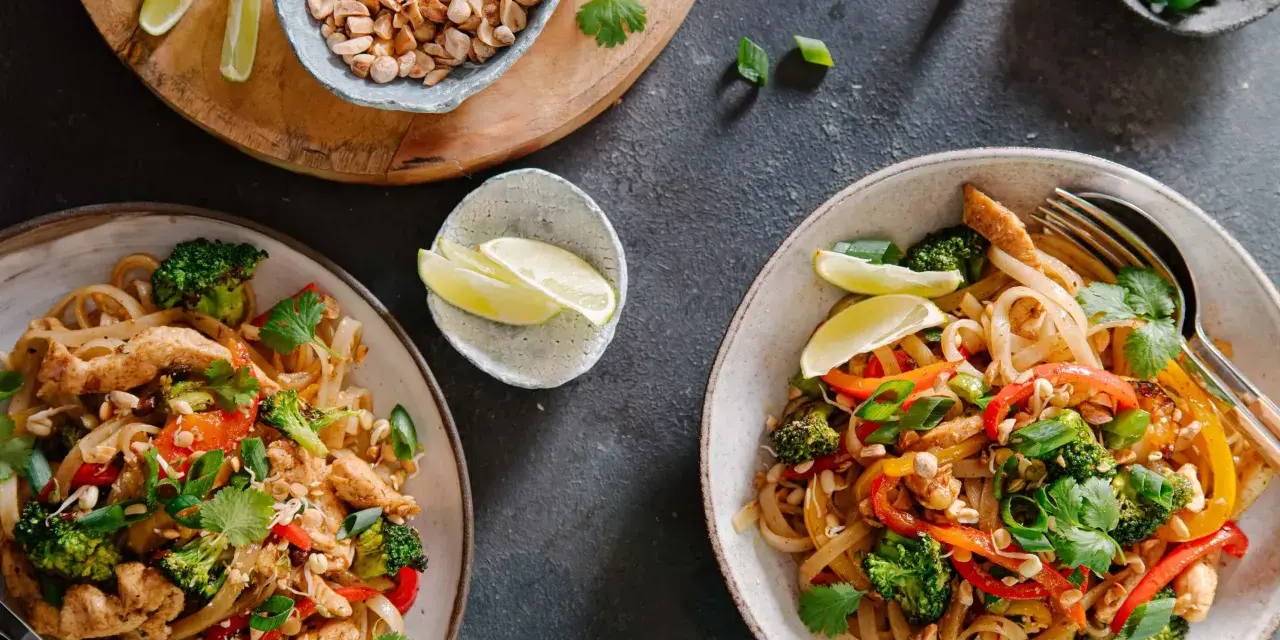 Dwa talerze z prostym pad thai z kurczakiem, brokułami, papryką i orzeszkami ziemnymi, podane z ćwiartkami limonki.