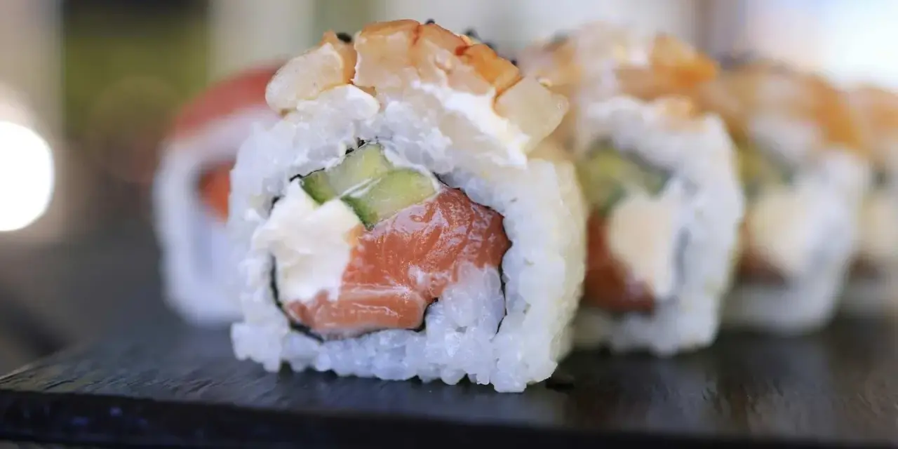 Uramaki co to? Rolka sushi z łososiem, awokado i krewetką, ryż na zewnątrz, na czarnej desce.