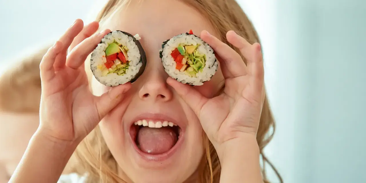 Uśmiechnięta dziewczynka trzyma kawałki sushi dla dzieci przed oczami, udając, że ma rolki zamiast oczu.