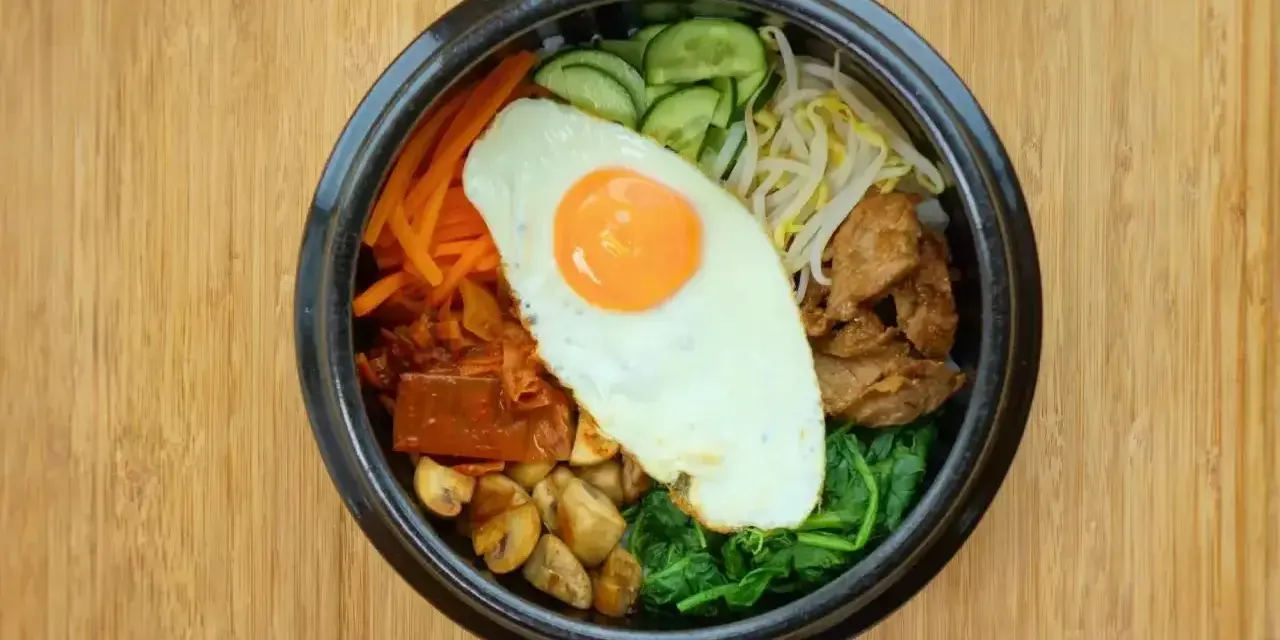 Apetyczne koreańskie jedzenie: miska bibimbap z jajkiem sadzonym, warzywami i mięsem na drewnianym tle.