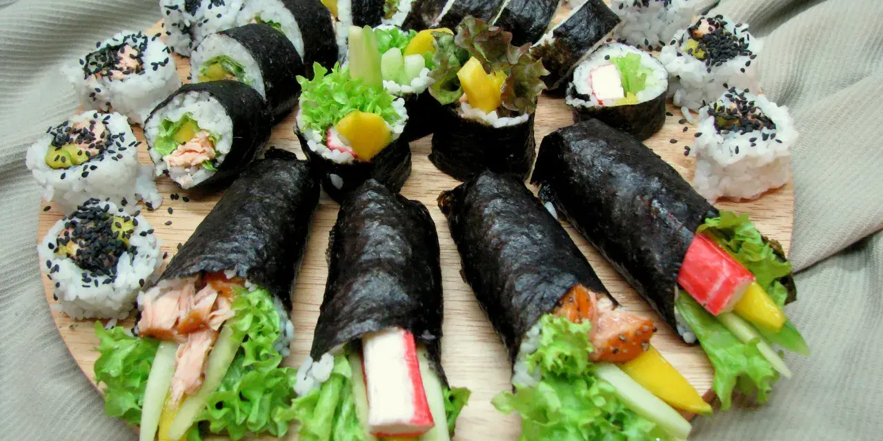 Talerz sushi z futomaki i uramaki. Widać rolki z łososiem, awokado, sałatą, paluszkami krabowymi i mango.