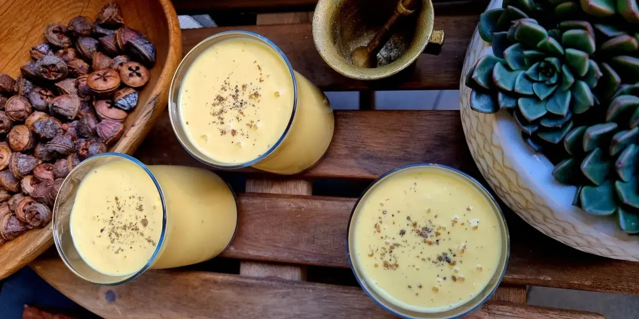 Trzy szklanki kremowego mango lassi, posypane przyprawami, obok miski z suszonymi owocami i sukulentu.