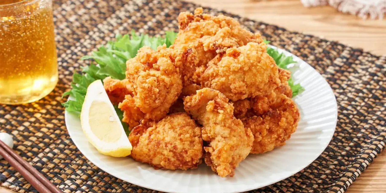 Chrupiące karaage chicken na talerzu, ozdobione plasterkiem cytryny i liśćmi sałaty. Obok szklanka złocistego napoju i pałeczki.
