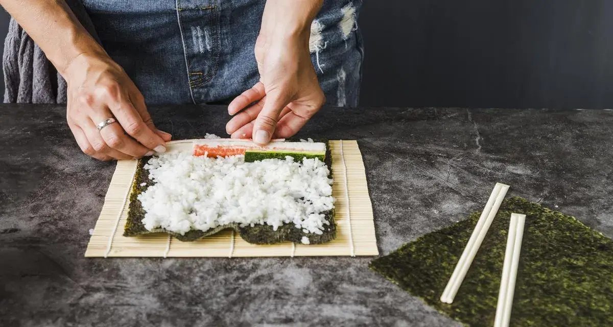 Ręce układają składniki do zwijania sushi na macie bambusowej. Ryż, paluszki krabowe i ogórek czekają na zawinięcie.