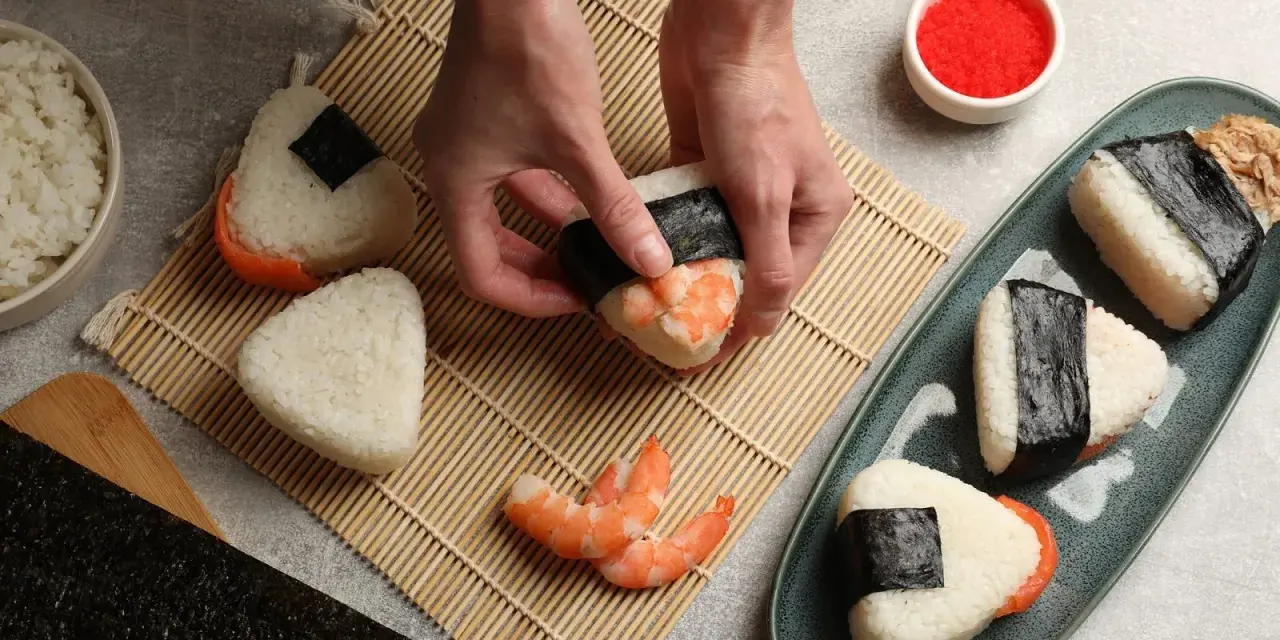 Ręce formują onigiri z krewetkami, tworząc pyszne japońskie jedzenie. Obok miska ryżu, nori i talerz gotowych przysmaków.
