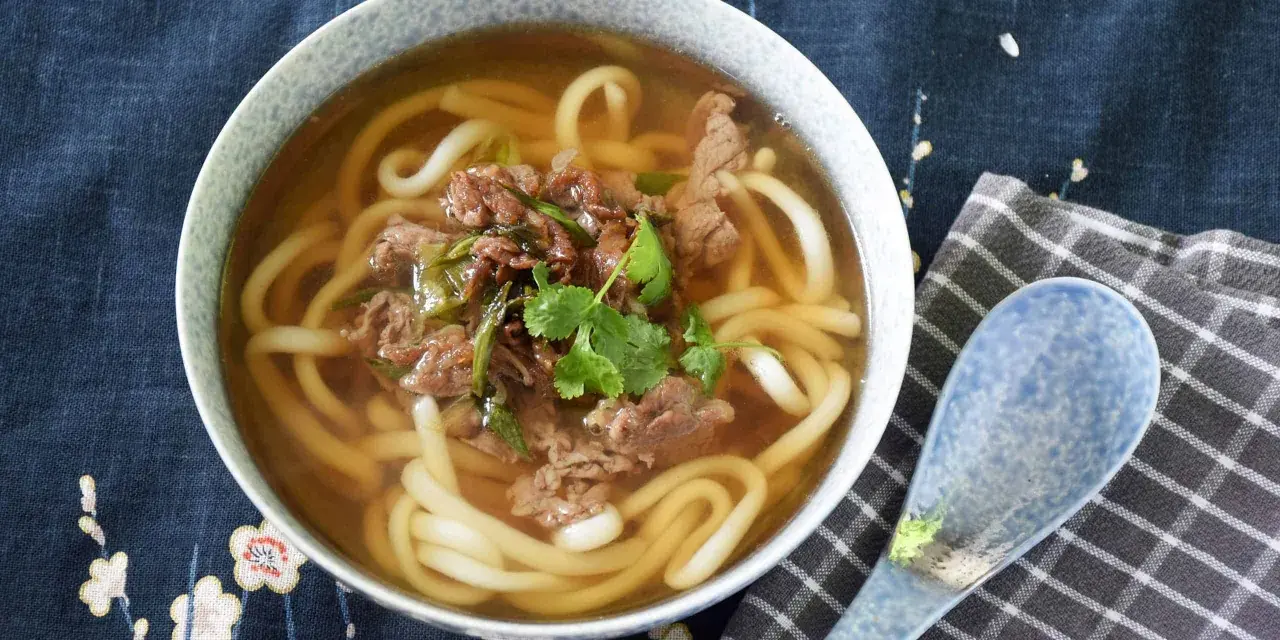 Gorąca zupa udon z wołowiną i szczypiorkiem, ozdobiona kolendrą, w misce na tle z motywem kwiatowym. Obok leży niebieska łyżka.