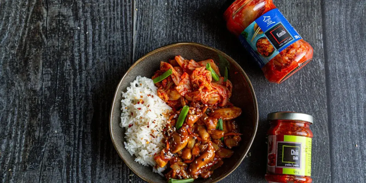 Miseczka ryżu z kurczakiem w sosie i kimchi, obok słoiczki z kimchi i pastą chili.