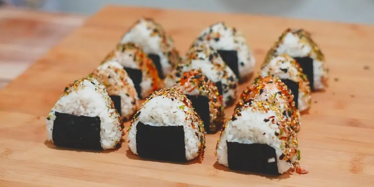 Rząd trójkątnych onigiri z kurczakiem, posypanych sezamem i przyprawami, ułożonych na drewnianej desce.